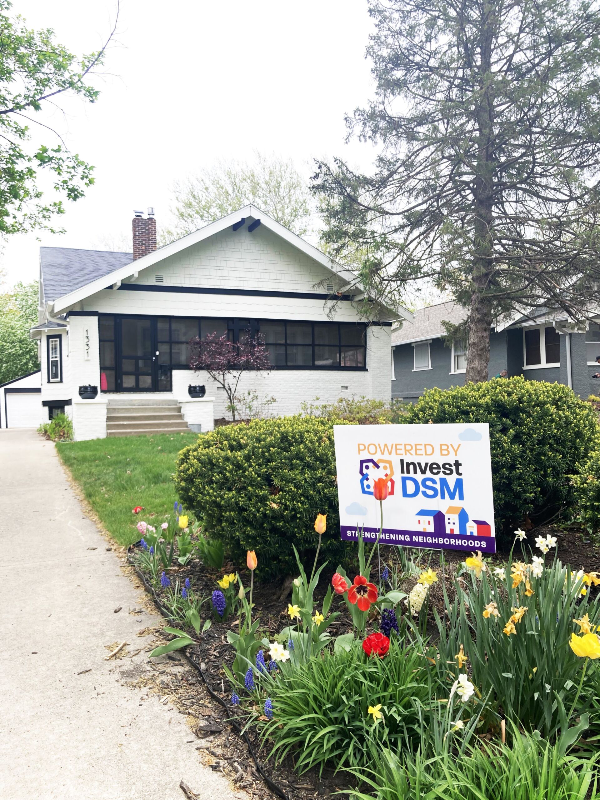 Invest DSM sign among flowers with home with updated white exterior in background